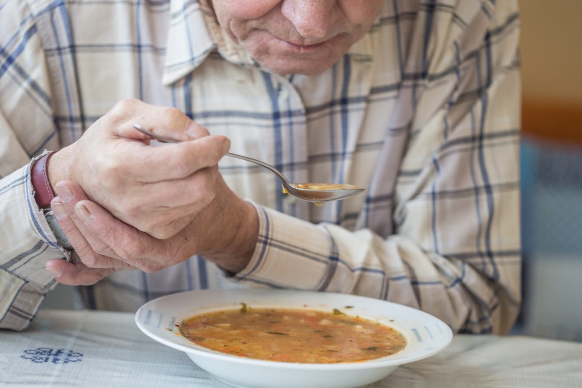 Mann spiser suppe og holder hånden mot skjelvinger ved Parkinsons sykdom