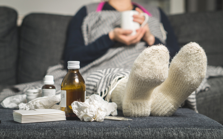 En syk kvinne ligger i sengen med varme sokker og piller og sirup ved siden av seg.