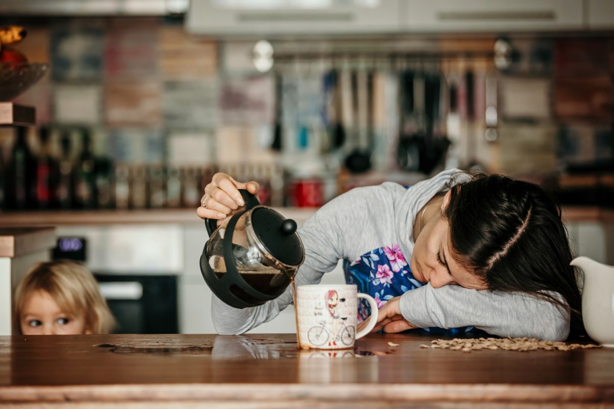 En sliten kvinne ligger på kjøkkenbenken og søler kaffe ved siden av et krus, og et barn står ved siden av henne.