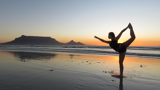 En kvinne praktiserer yoga på stranden. Solen har gått ned, men det er fortsatt lyst. Hun står på ett ben. Det er lavvann, og sanden ligger under et tynt lag med vann.