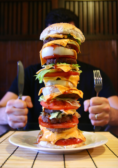 En mann sitter ved et bord med en høy hamburger på tallerkenen sin, en megaporce